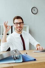 stylish smiling businessman with laptop and documents showing ok sign at workspace