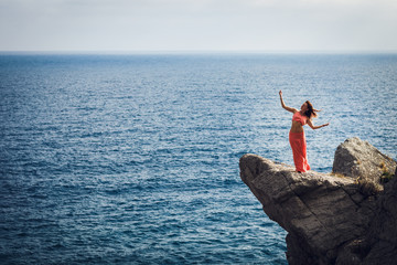 A beautiful young woman in hat with thin waist and charming forms dancing on a rock of high stone amid the blue sea on the ocean shore.The wind plays with long pink skirt and hair.All around  the sea.