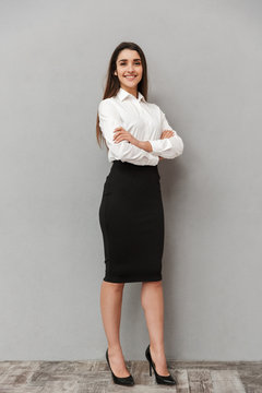 Full length portrait of caucasian woman with long brown hair in business wear smiling at camera and keeping arms crossed, isolated over white background