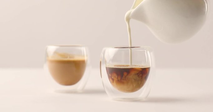 Pouring milk in one of two cups with coffee on white background