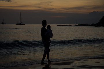 Image of Father carrying little baby girl in his arms. they are strolling and enjoying the beautiful sunset on the beach. Silhouettes of daddy and his little daughter at sea