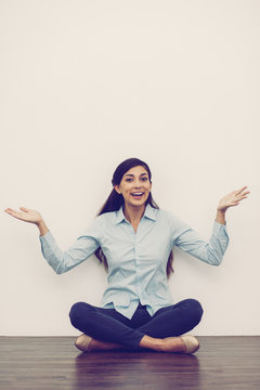 Portrait of smiling at camera middle-aged woman sitting on floor with crossed legs and outstretched hands with white wall in background. Front view.