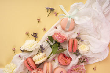 Pink and white macaroons cakes with big and small flower buds are decoratively laid on a white fabric on a yellow background