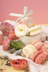 Pink and white macaroons cakes with big and small flower buds are decoratively laid on a white fabric and a cup on a yellow background
