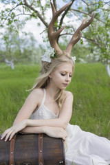 Naklejka premium Teen blonde girl wearing white dress with deer horns on her head, white butterfly and white flowers in her hair sits on the grass leaning on the ancient chest in a blooming garden