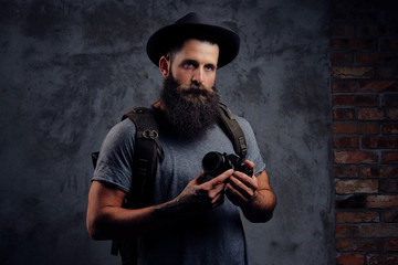 Portrait of a handsome bearded traveler in a hat with a backpack and tattooed arms, holds a photo camera, isolated on a dark background.