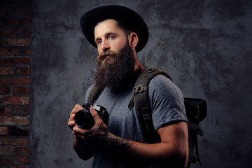 Portrait of a handsome bearded traveler in a hat with a backpack and tattooed arms, holds a photo camera, isolated on a dark background.