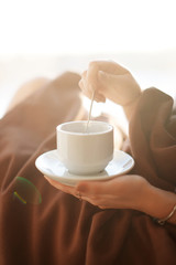 Young beautiful Girl drinking coffee sitting near the window in morning. Blond sexy naked female Boudoir photo standing in front of large window wrapped in blanket with her back to camera