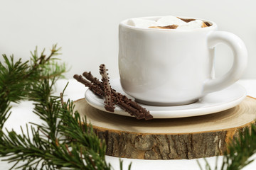 White cup of hot coffee with marshmallows and chocolate sticks on light background with Christmas tree twigs. Holiday concept. Selective soft focus.