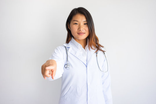 Smiling Asian Female Doctor Pointing At You.  She Is Looking At Camera. Medical Promotion Concept. Isolated Front View On White Background.