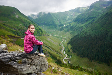 Fototapeta premium Smiling beautiful female looking to the camera sitting on rock in front of magnificent winding Transfagarashan road and green grassy mountains in Romania. Woman tourist climber happy spectacular view
