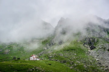 Beautiful white house standing isolated in low green grassy meadow near magnificent rocky mountains covered in fog. Amazing breathtaking nature view scenery landscape calm touristic