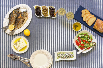 Freshly cooked seafood grilled sea bream fishes, sardines in olive oil and vegetable salad, olives, feta cheese, bread, water glasses served for two persons with copy space in the middle.