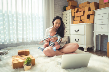 Fototapeta na wymiar Businesswoman with baby boy working at home using laptop and preparing package product on background. SME entrepreneur or freelance life style concept.