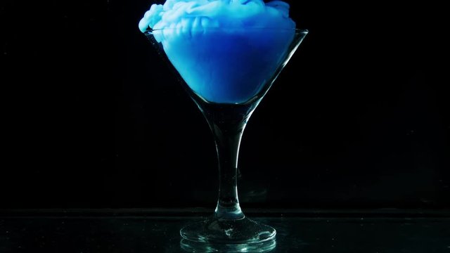 Beautiful glass goblet envelops thick blue smoke. A magical and magical potion is poured into a glass vessel on a black background. Slow motion.