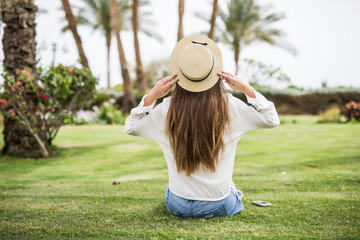 Fototapeta premium Rear view young beauty Woman in sunhat sitting on the grass on a beach under palms