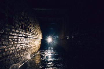 Man with flashlight in dark dirty brick underground tunnel or sewerage corridor - obrazy, fototapety, plakaty