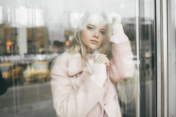 Unhappy pretty blonde girl is wearing pink coat behind the window, spring time