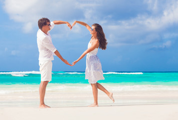 young happy couple in white making heart shape on tropical beach. honeymoon