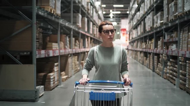 Hipster woman in glasses in store is buying goods. Beautiful woman is driving a shopping trolley in store. Girl do shopping