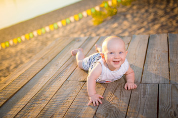 A child a boy, one year old, blond man lies on his stomach on wooden dock, pier in striped clothes, compound near pond on sandy beach against background of river in the summer at sunset of the day