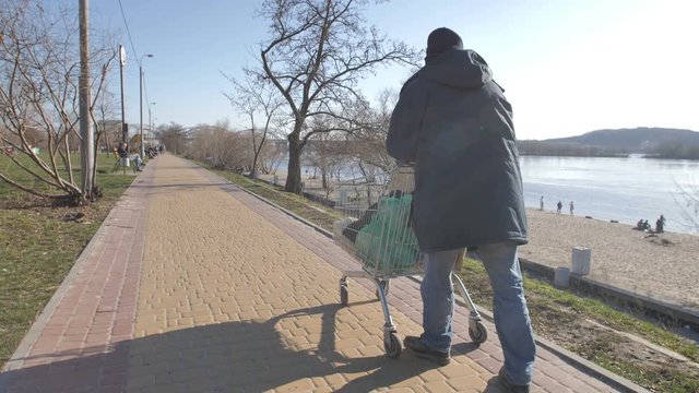 Back view of homeless man walking on the river bank in cold autumn pushing shopping cart with belongings. Desparate beggar male walking in city. Homelessness and social issues concept. Steadicam shot.