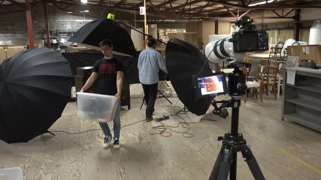 Photographer goes and adjusts the lights on the set while his assistant moves equipment.