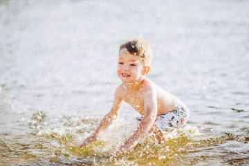 boy child playing makes splashes, beats hands on the water in the river at sunset of the day