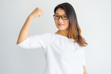 Powerful confident young Asian lady showing her muscle. Smiling successful woman in eyeglasses showing bicep and looking at camera. Strength concept