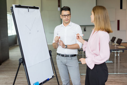 Confident Businessman And Businesswoman Discussing Strategy At Meeting. Young Caucasian Partners Or Colleagues Drawing Graph On Flipchart In Boardroom. Strategic Planning Concept