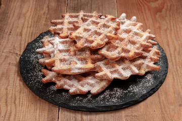 Wafers sprinkled with powdered sugar lie on a round, stone dish of slate. Slate lies on the background of wooden boards.