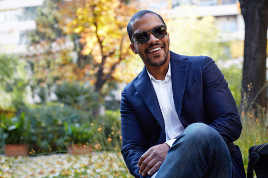 Portrait of Happy confident young African-American businessman in formal wear and sunglasses sitting at city park.Blurred background