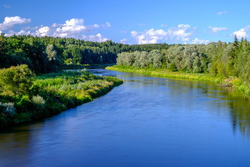 Bank of Gauja River in Latvia with blue sky and white clouds reflecting in water