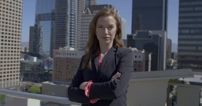 Medium shot, smartly dressed business woman with serious expression and folded arms, standing in front of skyscrapers in Downtown Los Angeles. Hand-held, slow motion 4K recorded at 60fps