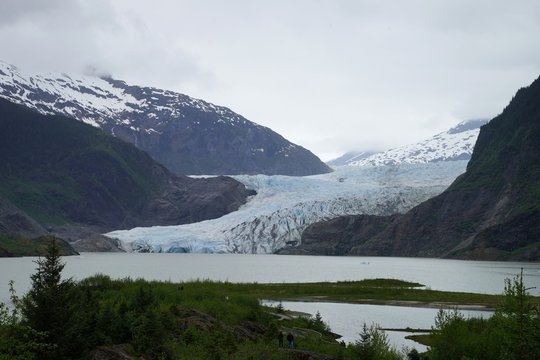 Blue ice of Mendenhall glacier with snow covered mountains on the background and hiking trail on the foreground