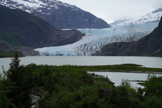 Blue ice of Mendenhall glacier with snow covered mountains on the background and hiking trail on the foreground