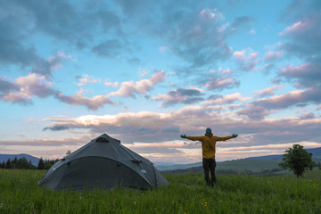 man standing next to a tent pitced on a meadow, looking at a clouds during sunset © listercz