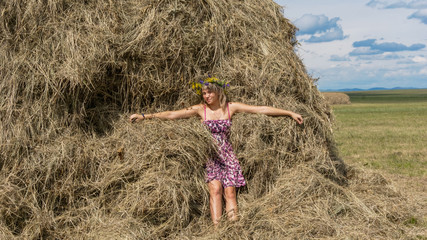 Naklejka na ściany i meble Pretty blondy girl wearing circlet of flowers lying on the haystack in the field, blue sky and haystacks background