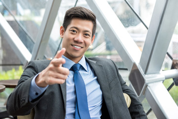 Smiling Asian businessman sitting in office lounge, pointing at camera