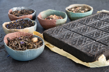 Brick of tea and various loose leaf teas in pottery bowls with a brass teapot