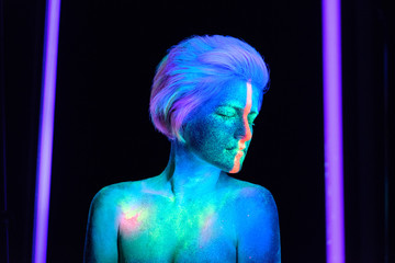 Model young beautiful girl portrait in studio with creative makeup, in neon ultraviolet lamp. Glows in the dark. Hair coloring with neon paints. Body art is blue, green, orange.