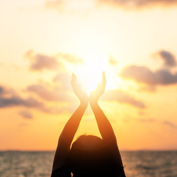 Summer sun June solstice concept and silhouette of happy young woman’s hands relaxing, meditating and holding sunset against warm golden hour sky on the beach with ocean or sea background