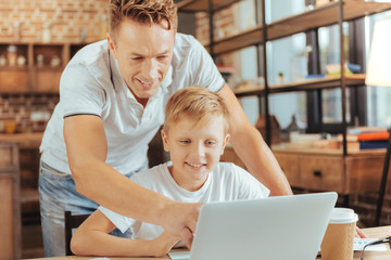 Happy fatherhood. Joyful positive nice man standing behind his son and looking at the laptop screen while pointing at the picture