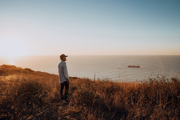 Man Standing on Coastal Cliff Overlooking Ocean at Sunset – Tranquil Outdoor Seascape with Golden...