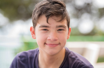 Close up portrait of a young teenager man looking at camera with a joyful smiling expression, against a green blur background. Outdoors portarits