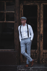 Fashionable retro dressed man with cap, suspenders and eyeglasses standing on city street and looking at side. Low angle view.