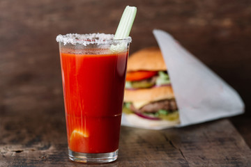 Glass of tomato juice with a celery stick and a big burger on a wooden background
