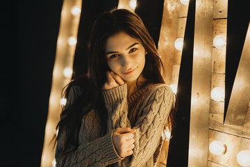 Portrait of beautiful young girl, sitting near a lights decoration and touching her face with one hand looking directly into camera.