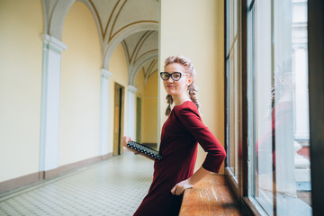 Romantic portrait of blond woman in eyeglasses wearing burgundy dress with book looking at you while standing near window sill indoor in hallway of old university building. Mood and study concept.