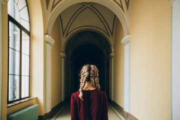 Naklejka premium Back view of romantic blond woman student with cute hairstyle two braids wearing burgundy dress while standing indoor in hallway of old university building. Mood and study concept.